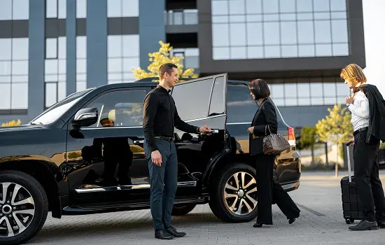 A chauffeur opens the door of a black SUV for business travellers with luggage outside an office building