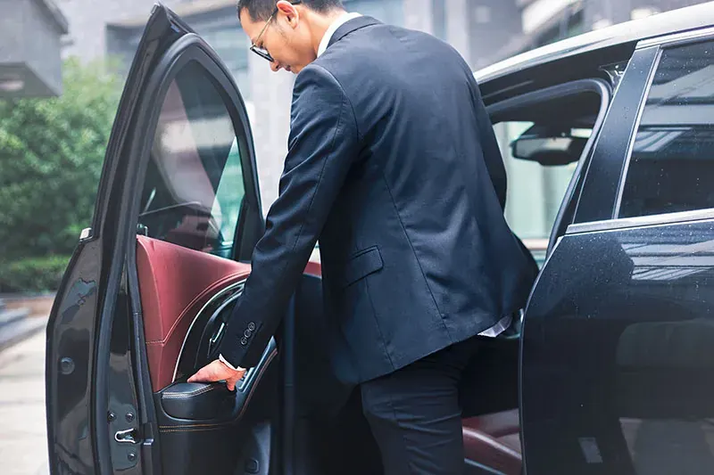 A chauffeur in a suit opens the rear door of a black car with red leather seats