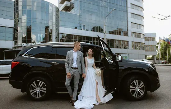 A bride and groom holding hands beside a black wedding car